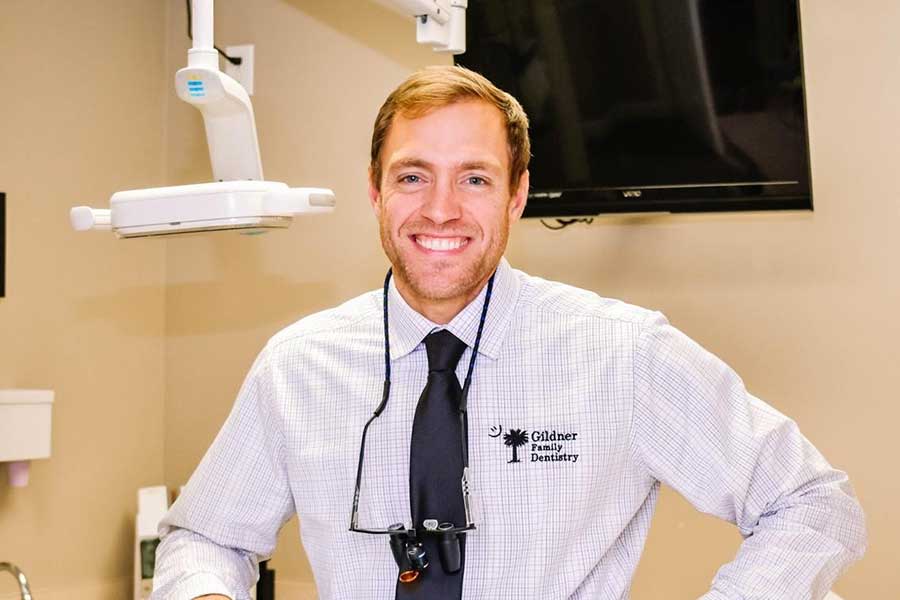 Dr. Robert Gildner, wearing a white shirt and black tie, smiling in a dental operation room under a monitor at Gildner Family Dentistry in Lexington, SC